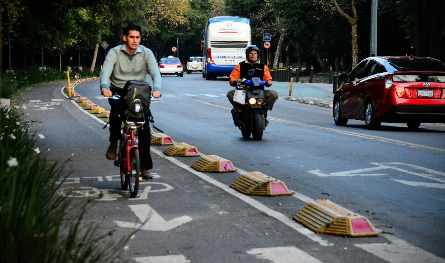 Ciclista circulando por una ciclovía protegida, en la calle circula de frente una motocicleta y dos vehículos a motor en sentido contrario