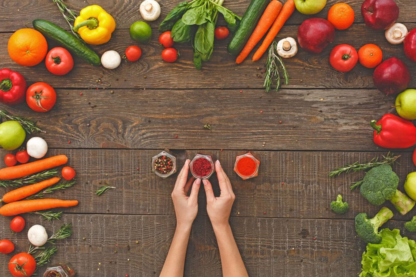 Foto de una mesa de madera vista desde arriba, con unas manos junto a tres frascos de especias, enmarcadas por un conjunto de verduras y hortalizas de diversos colores
