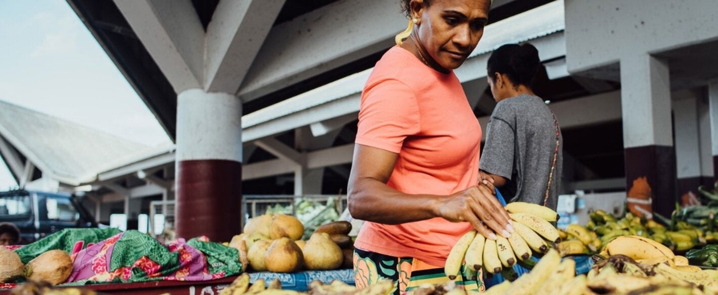 Photo of a middle aged woman dressed with an orange t-shirt grabbing a bunch of bananas on a market fruit stall