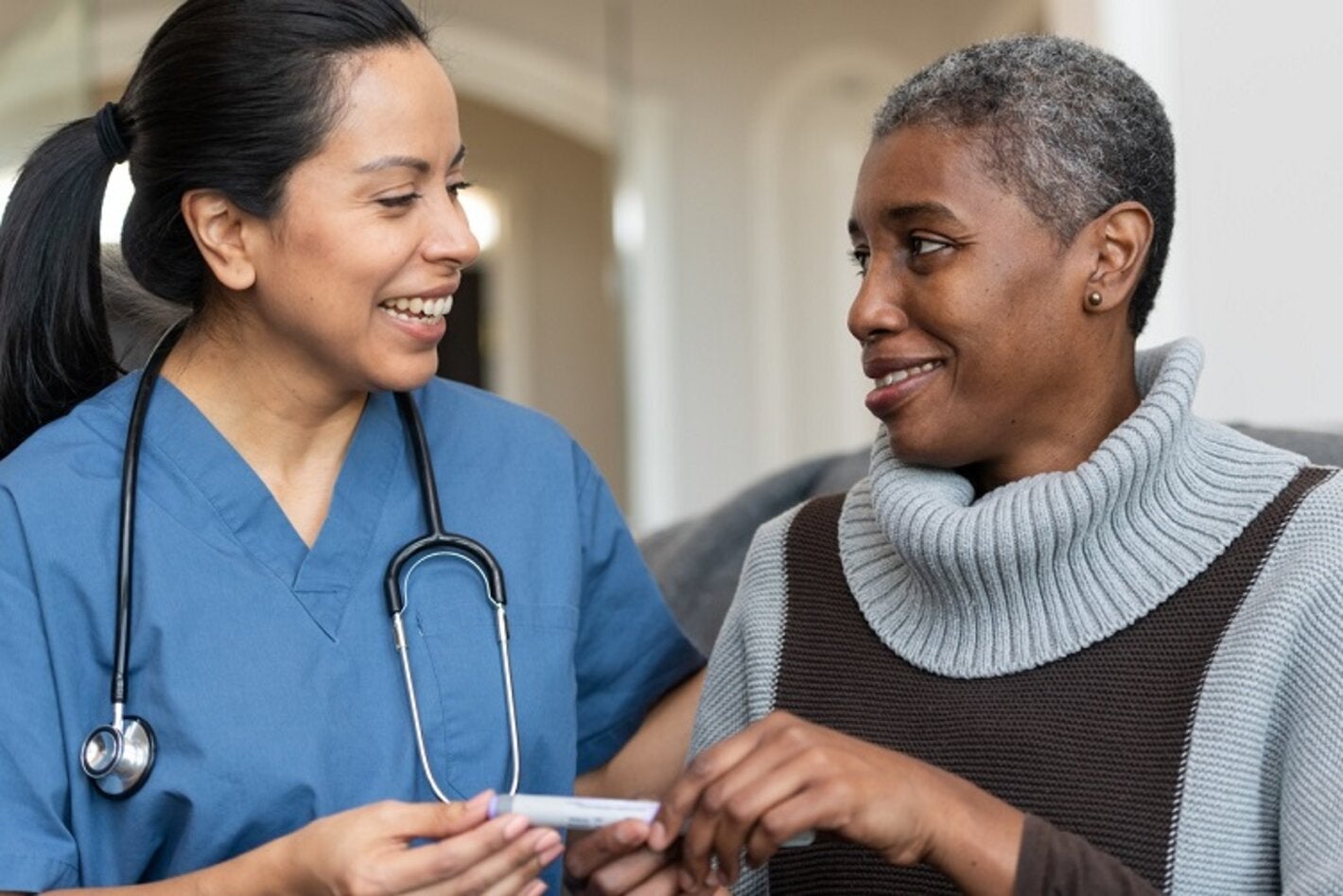 A senior African American female is being helped by her Hispanic female nurse during a home visit. They are listening to each other speak. She is showing her how to use an insulin pen.