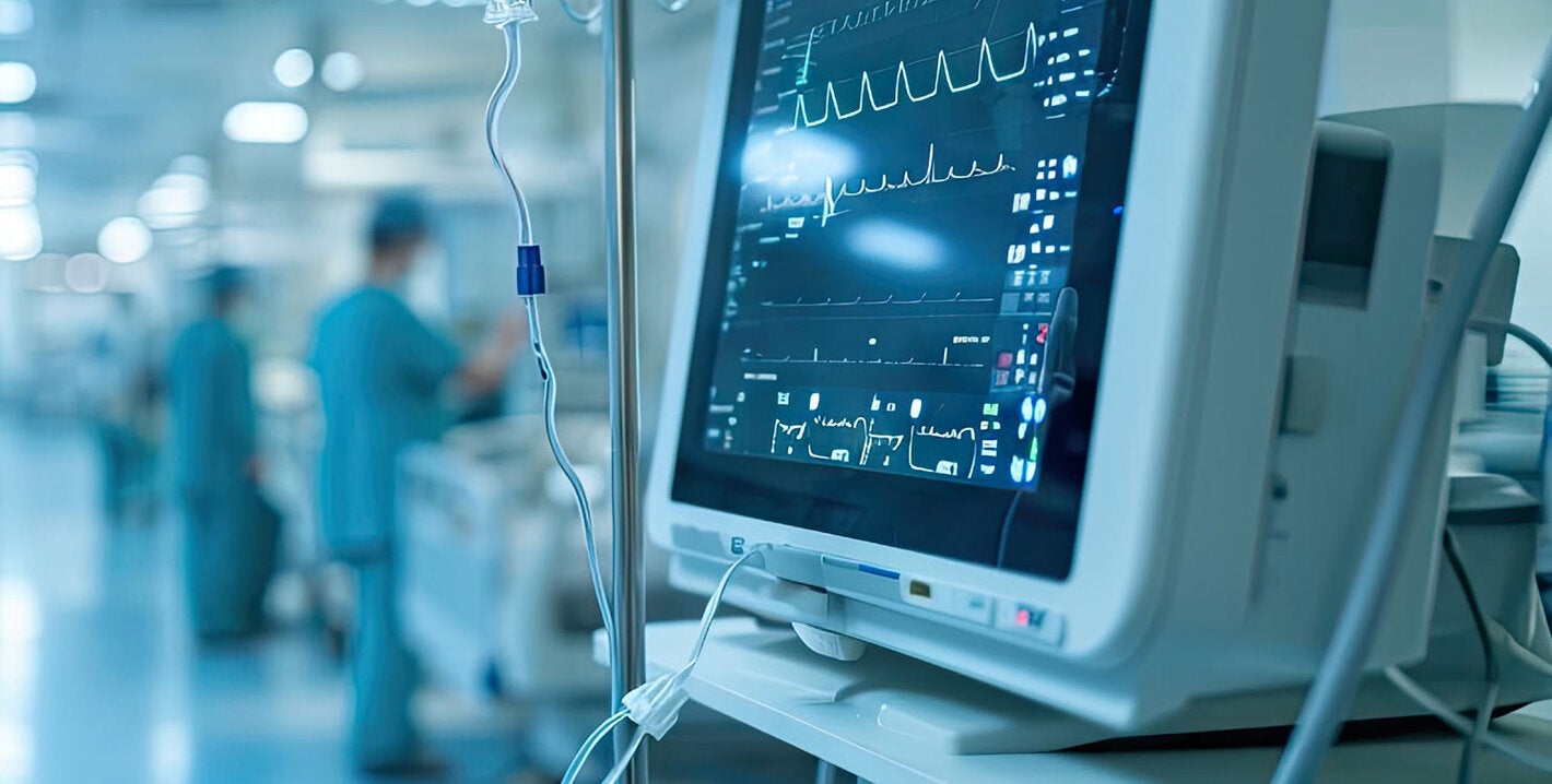 Hospital setting. The monitor with vital signs is in front. Behind are hospital beds and hospital staff.