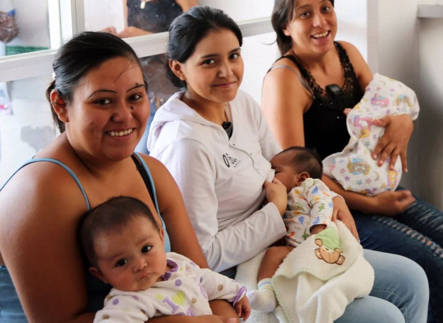 mujeres sentadas, mirando a cámara y sonriendo, con sus hijos bebés en brazos