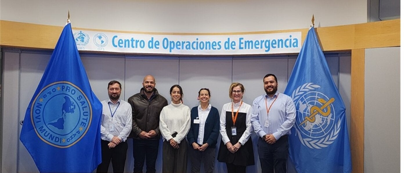Personas (hombres y mujeres) posan a la cámara en el Centro de Operaciones de Emergencias de la OPS. A ambos lados, están las banderas de la OPS y la OMS.