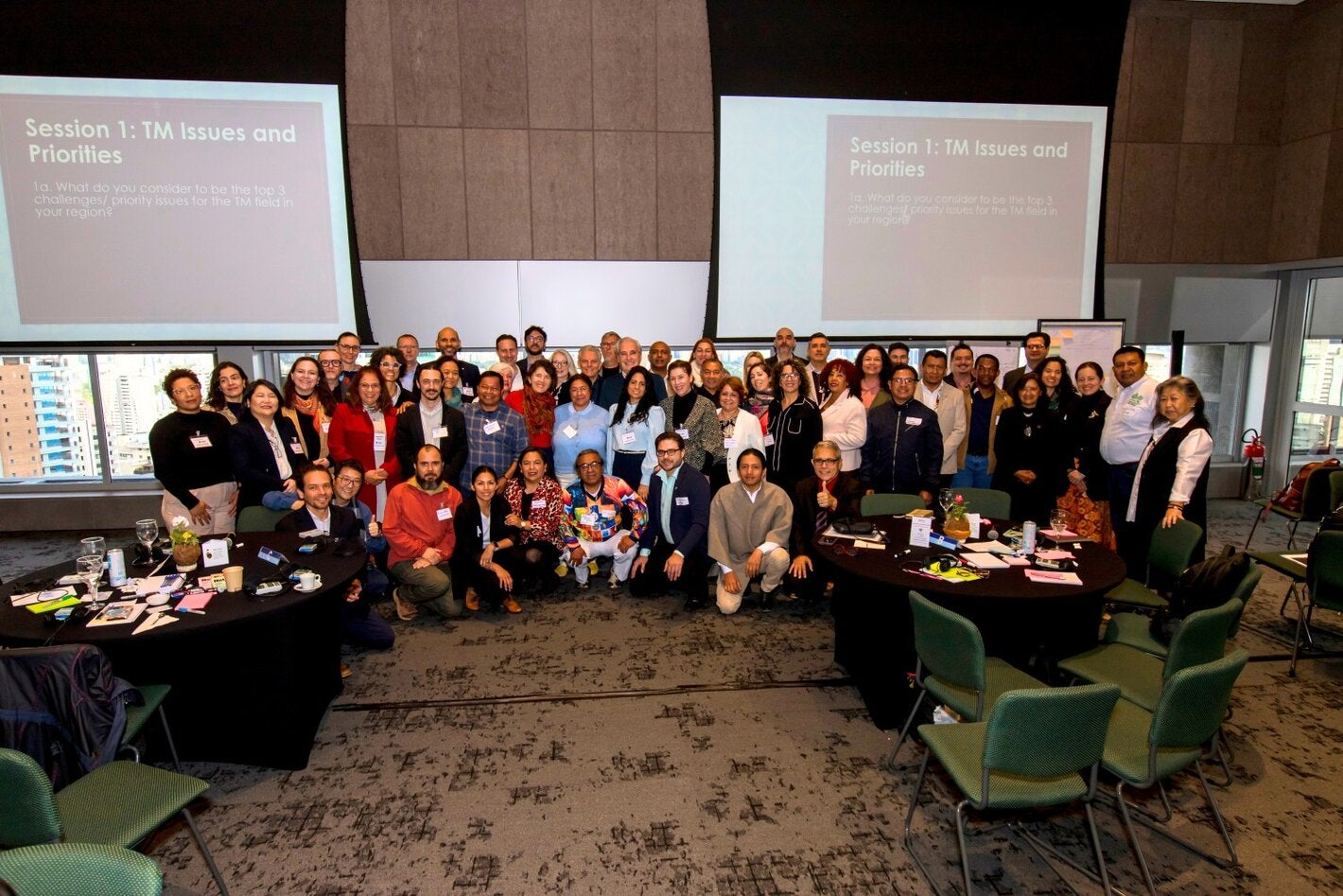 Group photo during the Regional Consultation for the Prioritization of TCIM Research in the Americas, held June 11-12, 2025, in São Paulo, Brazil. Approximately 50 participants pose smiling in the center of a room with tables and working materials. In the background, two screens show the agenda for the “TM Themes and Priorities” session.