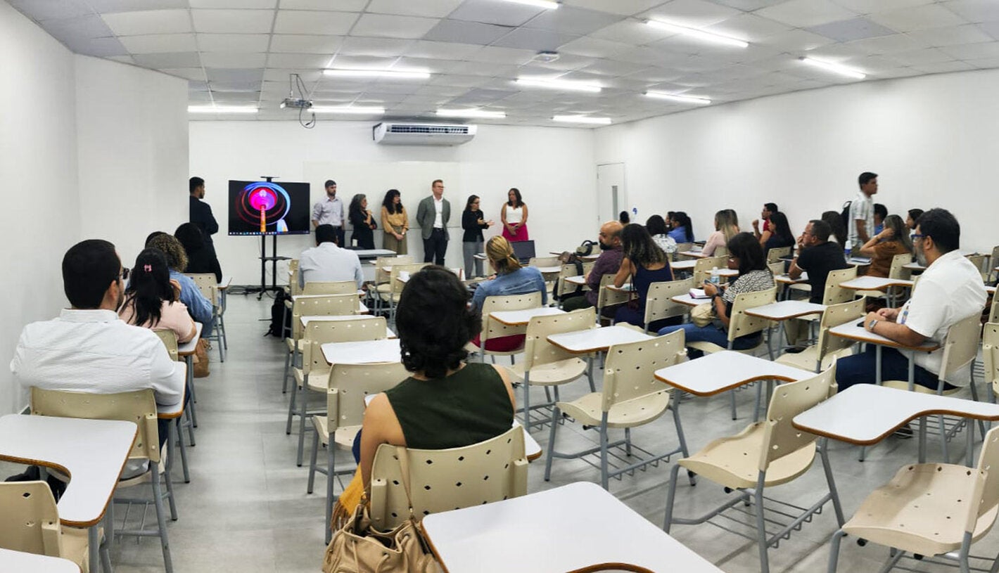 Profissionais sentados em mesas de sala de aula, ouvindo palestra da equipe da OPAS e parceiros