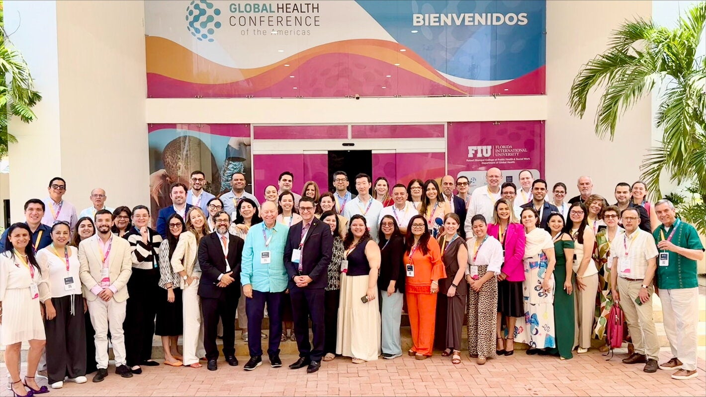 Los participantes del taller “Evidencia y herramientas disponibles para la eliminación del cáncer cervicouterino en las Américas” posan para una foto grupal frente a la entrada de un edificio. En el fondo, se puede ver un cartel que dice "GLOBAL HEALTH CONFERENCE of the Americas" y "BIENVENIDOS". También hay logotipos de FIU (Florida International University) y otros elementos decorativos. Los asistentes están vestidos con ropa formal y llevan gafetes colgados al cuello.