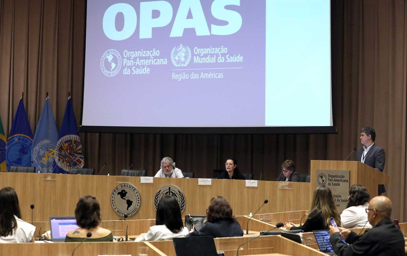 Mesa de abertura do evento, com telão ao fundo com a logo da OPAS