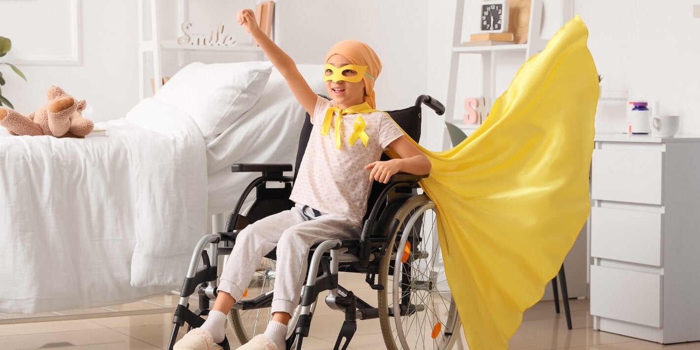 A child sitting in a wheelchair in a bright bedroom raises one arm in a triumphant pose, wearing a yellow cape and awareness ribbon, symbolizing strength, hope, and resilience during childhood cancer treatment.