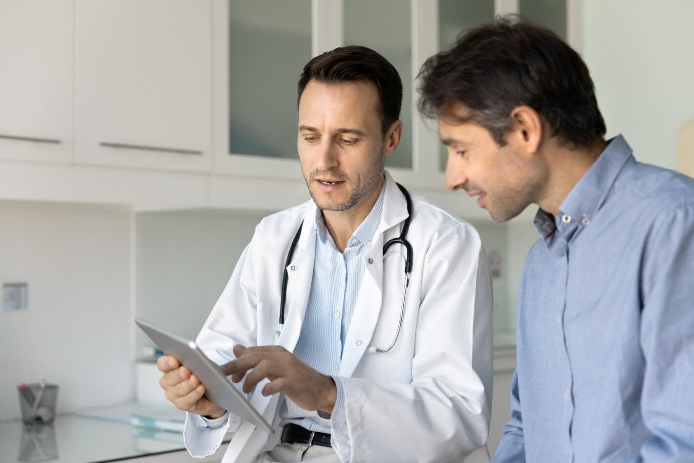 Male doctor with a white coat shows a clipboard to a male patient dressed with a blue shirt.