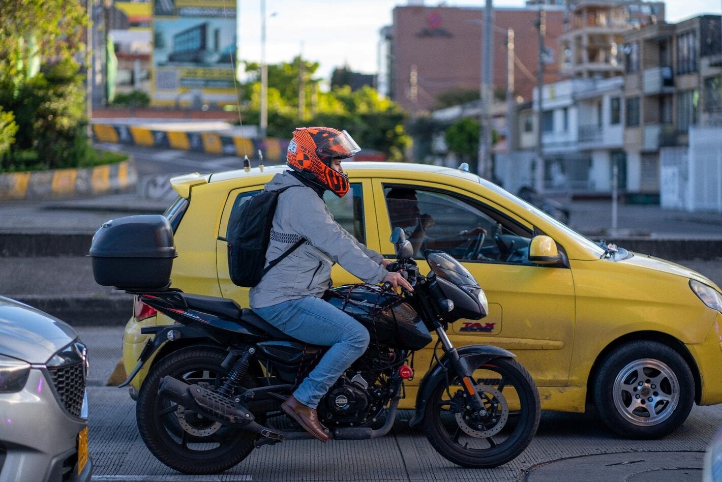 Motociclista con casco rojo, circulando en una avenida urbana, con un vehículo amarillo a su lado izquierdo