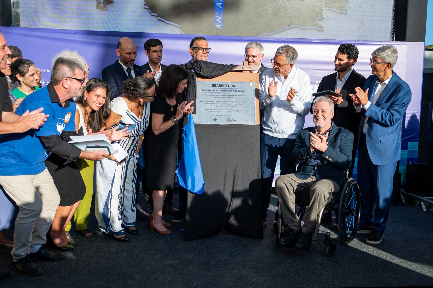 Grupo de autoridades, pesquisadores e participantes reunidos ao redor da placa de inauguração durante a cerimônia de reabertura do Centro Cultural do Ministério da Saúde, no Rio de Janeiro. Algumas pessoas aplaudem enquanto a placa comemorativa é descerrada; em primeiro plano, um homem em cadeira de rodas sorri e participa do momento.