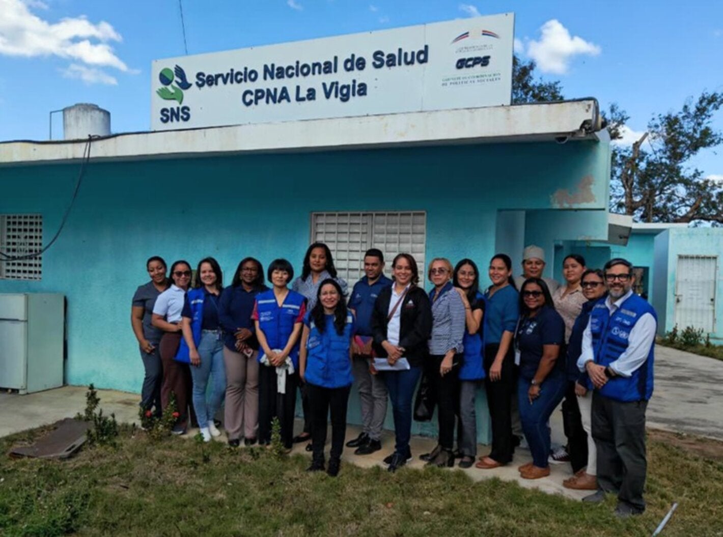 Grupo de trabajadores de salud del centro de atención primaria La Vigía y expertos de OPS posan frente al centro de salud, que tiene las paredes pintadas de verde aguamarina, con un cielo azul con nubes dispersas y un frente de hierba