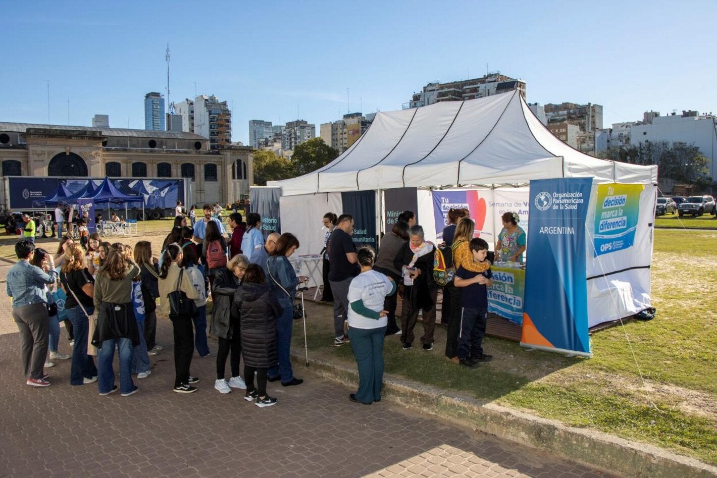 Un grupo de personas espera frente a un gazebo blanco, con banderas y banners de OPS, para aplicarse sus vacunas en la Feria del Libro.