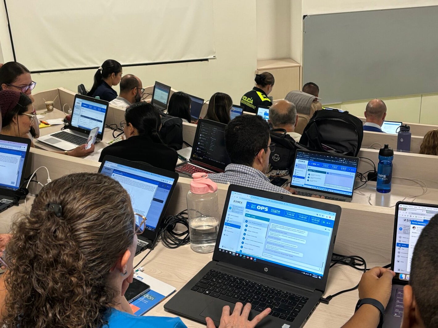 personas en salón de clases tipo auditorio, usando computador. Plataforma SIMEX visible en la pantalla.