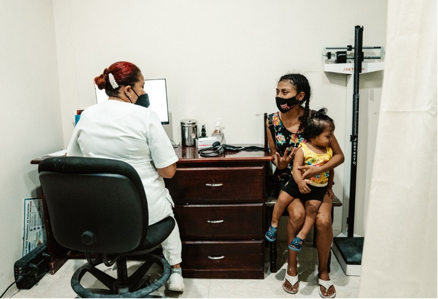 Personal de salud sentado en un consultorio conversa con una persona adulta que sostiene a una niña pequeña en brazos.
