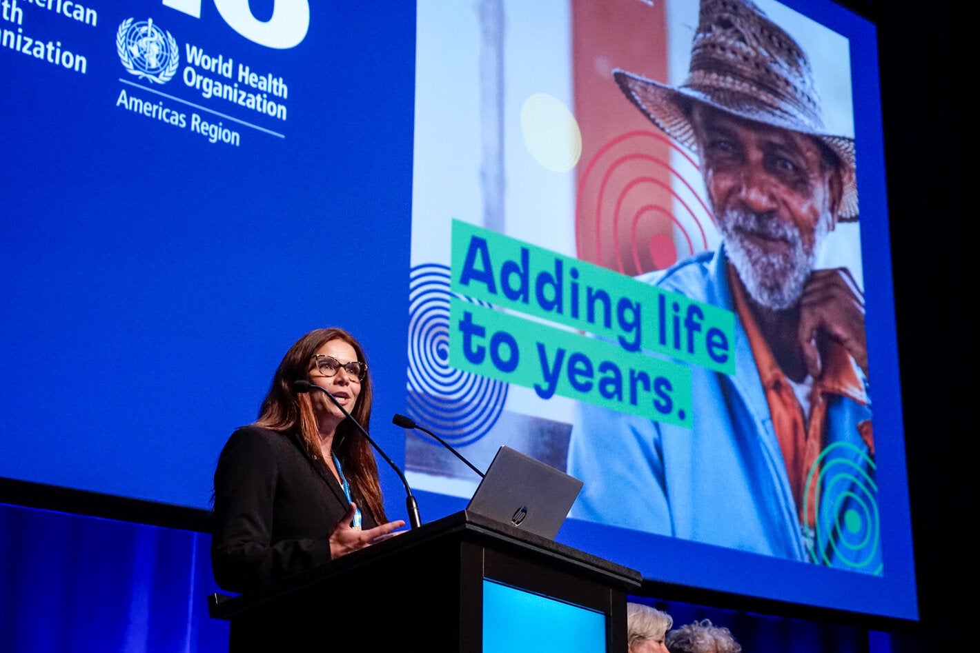 Woman on podium giving speech with presentation on a big screen behind her, with the Decade of Healthy Aging in the Americas card with slogan that reads "Adding life to years"