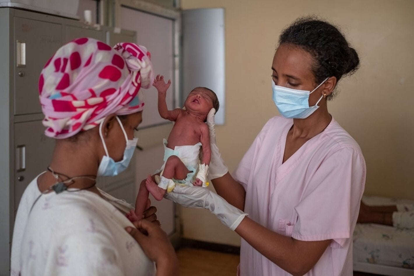 doctor with mask holding newborn baby in clinic in front of its mother