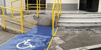 Rampa accesible para sillas de ruedas con pasamanos amarillos que conducen a un hospital. Un símbolo de discapacidad azul y blanco está pintado en el suelo.