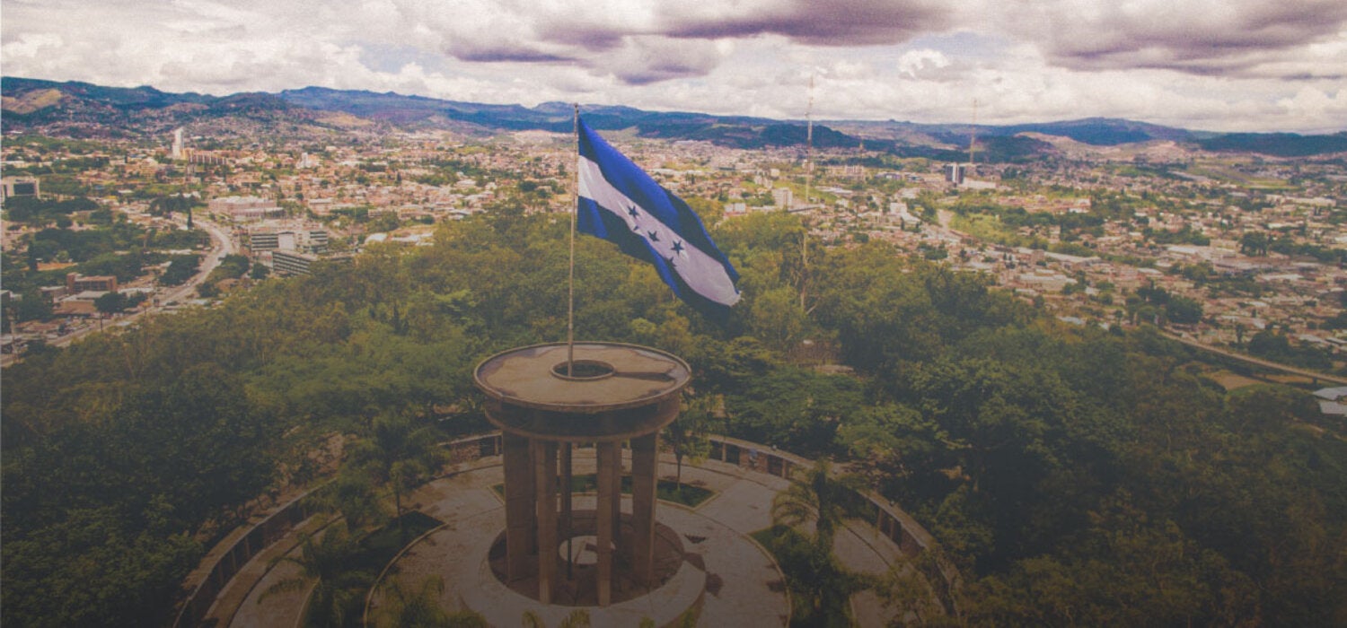 Foto de un paisaje con la bandera de Honduras en primer plano ondeando desde lo alto de una colina, montada sobre una columna circular. Al fondo, panorámica de la ciudad de Tegucigalpa y en el horizonte una línea azul de montañas