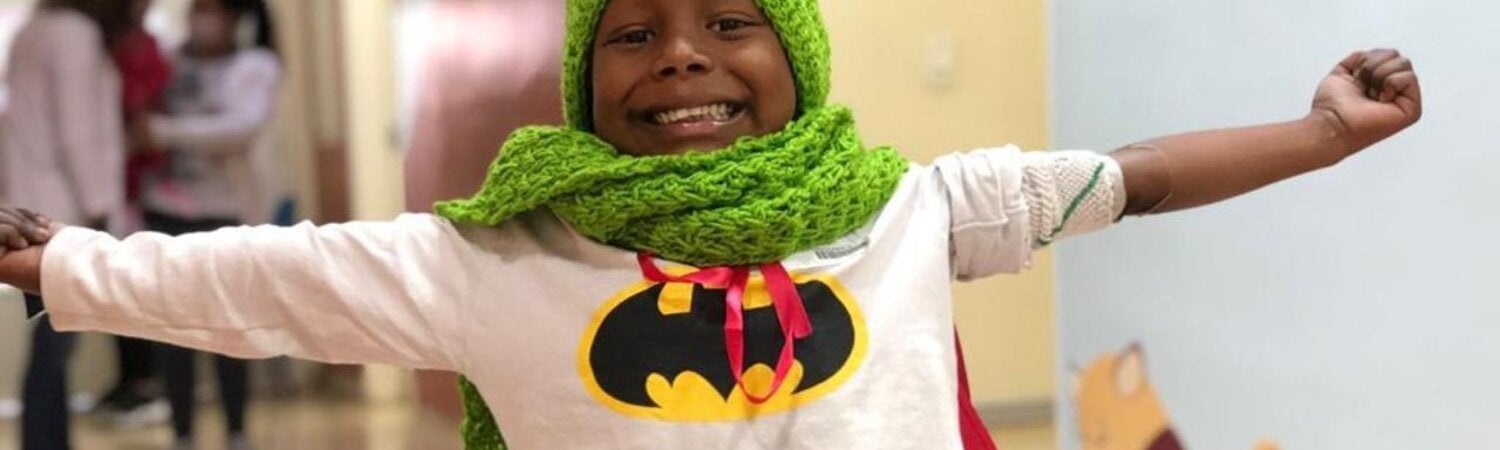 Niño afrodescendiente con los brazos extendidos y una gran sonrisa en su rostro. Vestido con una camiseta con el logo de Batman y un gorro y bufanda tejidos con lana verde