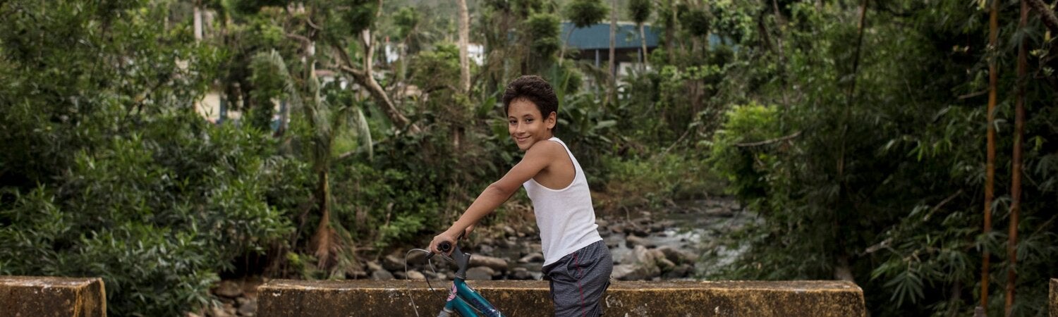 A boy of around 10-years-old is seated on a bicycle with his feet on the ground and his hands holding the bike, over a cement bridge. In the background, a forest of tropical trees and bushes hide the roofs of a building ant the shilouette of a mountain