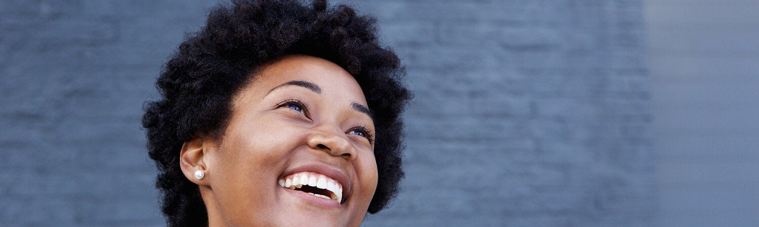 Fotografía de una mujer afrodescendiente, de pelo corto, sonriendo con la mirada hacia arriba. Detrás un fondo de ladrillo gris