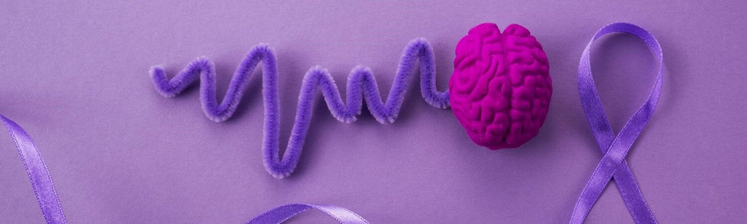 Sobre fondo morado, una ilustración de un cerebro en rosa fucsia del que sale una línea morada hacia la izquierda, al lado derecho un lazo de tela morado