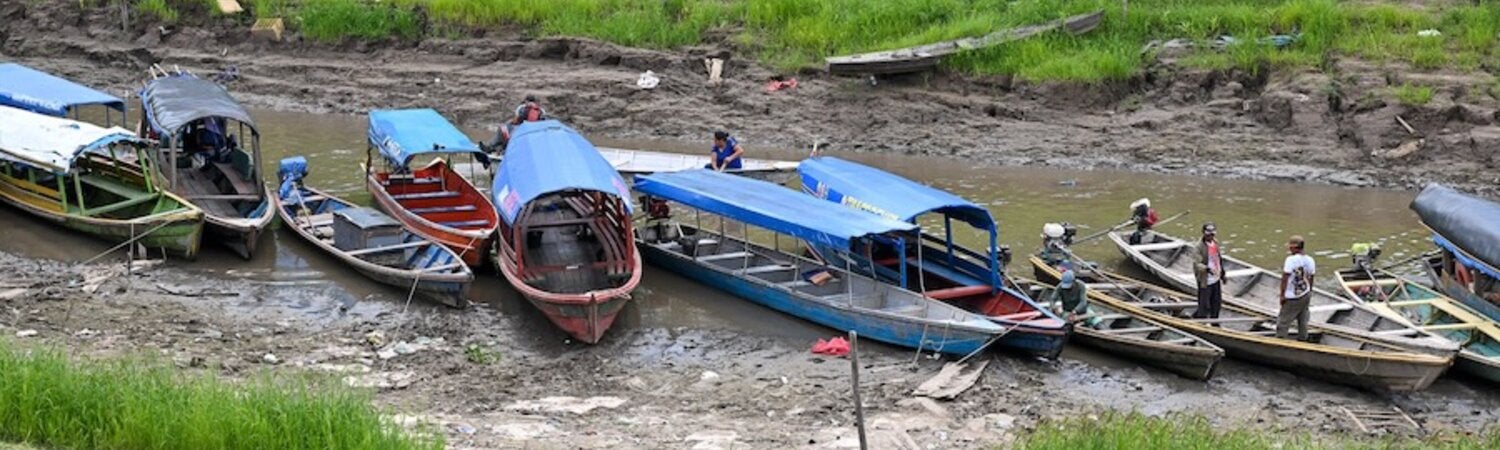 Wooden boats moored in a shallow waterway, with exposed riverbanks and low water levels visible along the channel during a period of reduced river flow in the Amazon region.