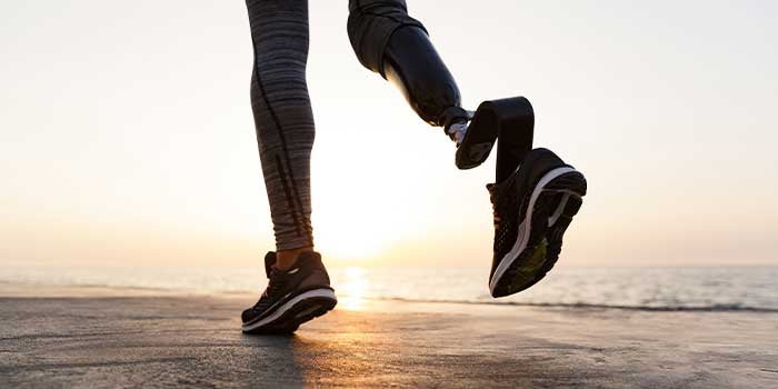 A women is in running shoes and leggings running on a beach towards a setting sun. The photo is focussed on the back of her two legs, one of which is a prosthesis